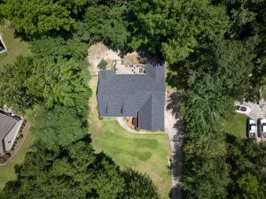 Wide angle aerial image of a residential roof with granite black aphalt shingles. Home surrounded by green grass and full green trees.